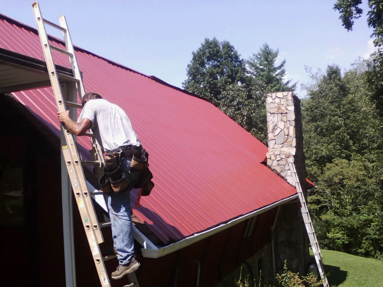 Roofer on a ladder beside a red metal roof and stone chimney — Soffit & Fascia Repair by Roofing Brothers in Hammondsville